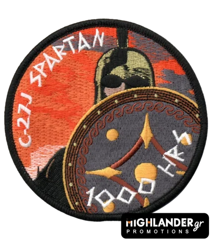 Στρογγυλό κεντητό σήμα με πορτοκαλί φόντο δύσης. Απεικονίζει Σπαρτιάτη πολεμιστή με ασπίδα και τα κείμενα "C-27J SPARTAN" και "1000 HRS".