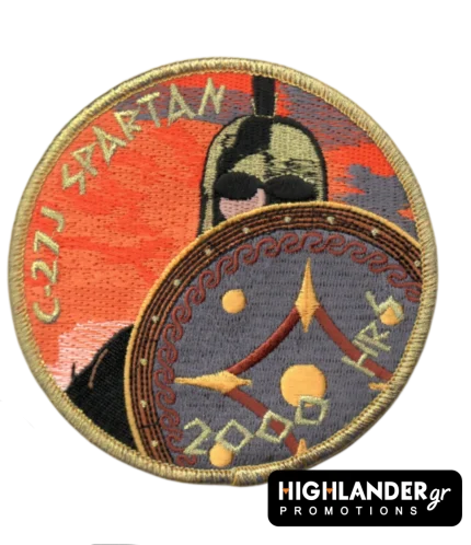 Στρογγυλό κεντητό σήμα με χρυσό περιθώριο. Απεικονίζει Σπαρτιάτη πολεμιστή με ασπίδα σε πορτοκαλί φόντο και χρυσά γράμματα "C-27J SPARTAN" και "2000 HRS".