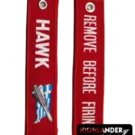 Red fabric keychain with embroidered HAWK missile and Greek flag on one side and white "REMOVE BEFORE FIRING" text on the other.